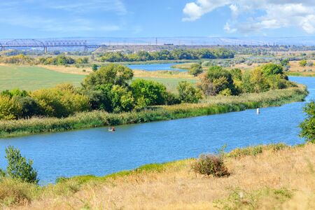 The wide bend of the Southern Bug River is immersed in the greenery of the garden and dried grass on the slope in the foreground, the river reaches the railway bridge on the horizon.の写真素材