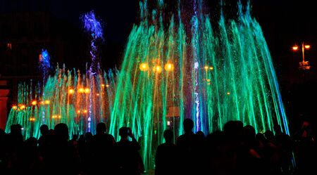 Dark silhouettes of people around the fountainin the late evening, multi-colored neon lights illuminate the jets of a powerful urban singing fountain.の写真素材