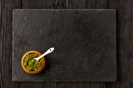 Matcha green tea powder in oak cup with white ceramic spoon with kitchen cutting board made of black slate, on wooden dark background, flat lay, copy space.の写真素材