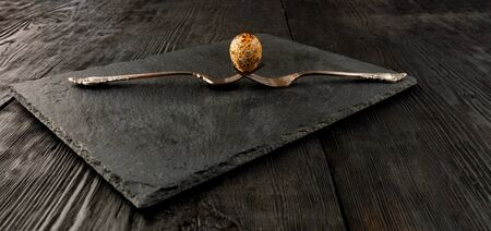 Quail egg on two old crossed forks on a kitchen cutting board in black of slate and a dark wooden...