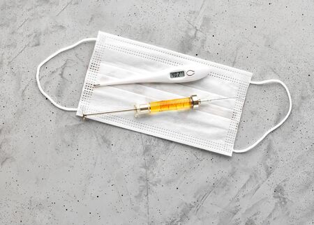 Medical mask, electronic thermometer and syringe with a yellow vaccine on a gray concrete...