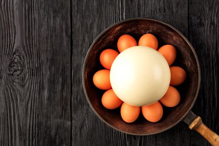 A large ostrich egg surrounded by chicken eggs in an old cast-iron pan that stands on an old black wooden surface, low key image, top view, copy space.の写真素材