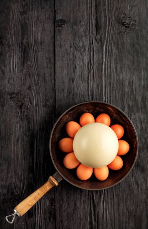Ostrich egg, selective focuse, surrounded by chicken eggs in an old cast-iron skillet, which is standing on an old black wooden surface, top view, copy space, vertical image.の写真素材