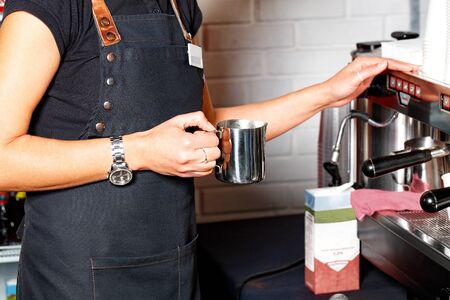 The barista in a black apron holds in his hand a metal mug for making coffee latte on the background of a coffee machine and a packet of cream in blur.の写真素材