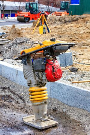 A gasoline vibratory rammer stands on a mixture of rubble and sand against the background of a road...