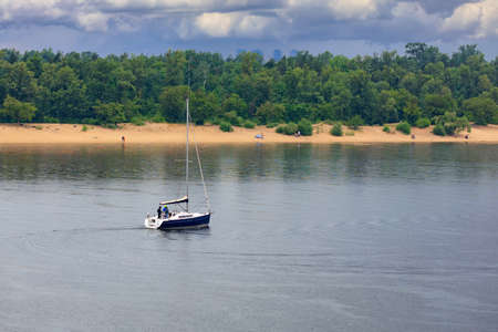 Sailing boat with a bare mast swinging on the river waves in the rain against the background of the river bank and cloudy rainy sky. Light in a dramatic gloomy sky.の写真素材