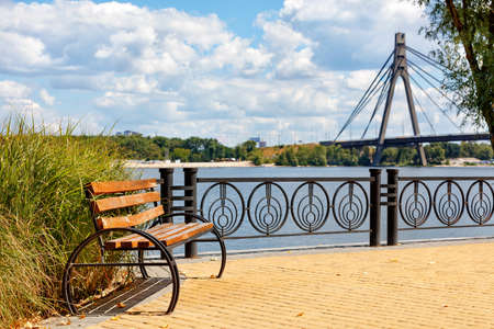 A beautiful wooden bench on the Dnipr? embankment in Kyiv. Sidewalk yellow tiles against the backdrop of sunlight on a bright summer day and the blurred north bridge over the river.の写真素材