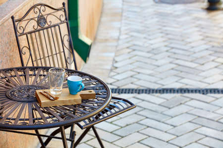 Street still life of a stylish metal table with a turquoise cup, a glass with a metal spoon on a wooden stand, against a background of paving stones in blur, selective focus, text space.の写真素材