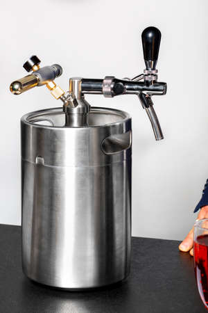 A new clean stainless steel barrel and a tap on it, with a glass of red wine and part of a man's hand visible in the foreground in the corner.の写真素材