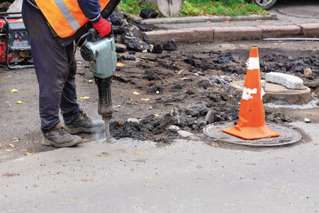 A road worker, dressed in reflective clothing, uses an electric jackhammer to rip old asphalt off the road and uses a gasoline generator, copy space.の写真素材