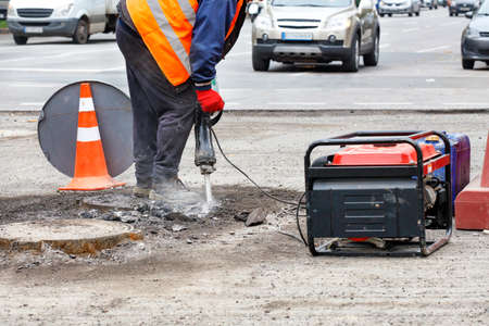 A road worker, dressed in reflective clothing, repairs a section of road near sewers with an...