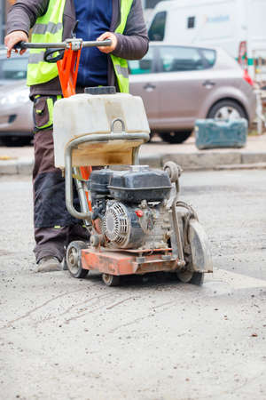 A road worker in reflective clothing cuts old asphalt on the road with a gasoline cutter against the...