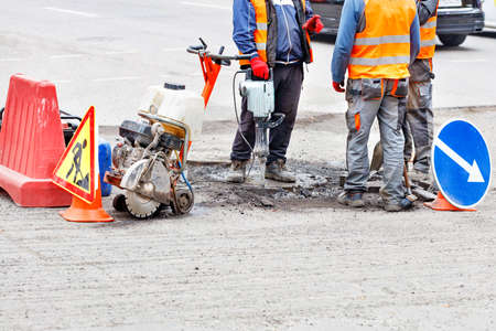 Road workers in reflective clothing on a fenced-in stretch of road discuss a roadway repair plan using an electric jackhammer, petrol cutter, shovel and brush. Copy space.の写真素材