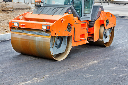 The metal cylinders of the large vibratory roller roll over the new road surface for powerful...