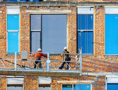 Builders install large windows on the facade of a house under construction using a cradle and an aluminum profile, which is a concept for saving heat in the house.の写真素材