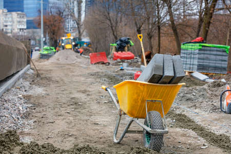 Construction yellow wheelbarrow and curb concrete afternoon blocks at the sidewalk repair construction site against the background of a city street on a spring in blur. Copy space.の写真素材
