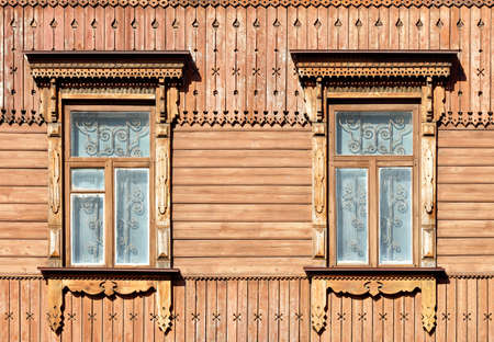 The old wooden textured facade of the house with carved platbands, curly wall elements and metal bars in the windows, illuminated by warm sunlight.の写真素材