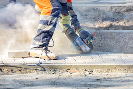 A builder cuts paving slabs with an electric grinder and diamond cutting disc, creating a cloud of...