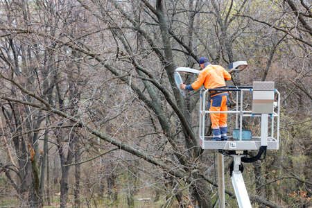 A utility worker in an orange uniform uses a construction hoist to repair street lighting on a pole...