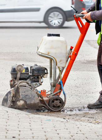 A road worker in a reflective vest cuts old asphalt on the road in front of a city street with a...