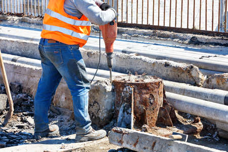 A road worker in an orange reflective vest works with a jackhammer to smash an old reinforced concrete structure around a rusty support. Copy space.の写真素材