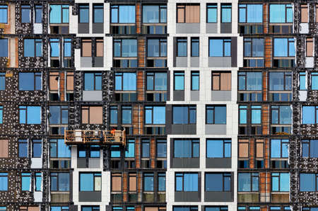 Fragment of the facade of a multi-storey residential building under construction at the stage of its insulation with mineral insulation and sheathing with decorative panels using a winch and a mobile platform.の写真素材