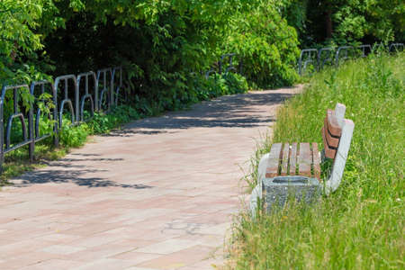 On a summer afternoon, an empty park bench sits along a cobbled footpath among lush green...