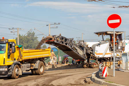 A crawler milling machine removes the top layer of asphalt from a road section being repaired and loads it into a dump truck in front of a city street on a summer day.の写真素材