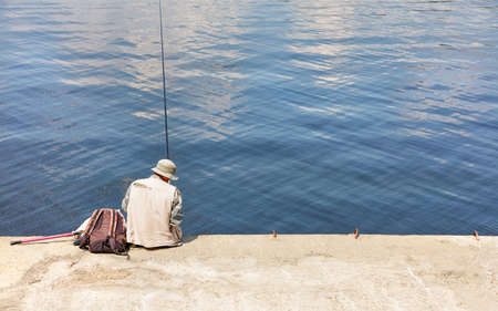 A fisherman in a light-colored panama hat and a vest sits on a concrete pier with a fishing rod near the water on a bright sunny day. Copy space.の写真素材