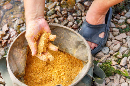 A fisherman prepares fish bait for fishing in a tarpaulin bucket against the background of a pebble river bank in a blurred form and sunlight. Selective focus, close-up.の写真素材