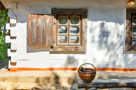 A wicker basket with an clay jug stands in the courtyard on a wooden bench near an old window with wooden shutters of a Ukrainian rural hut under a thatched roof in the sun.の写真素材