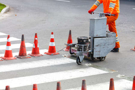 An orange overalls road worker renews pedestrian crosswalks on a road using a power trolley and...