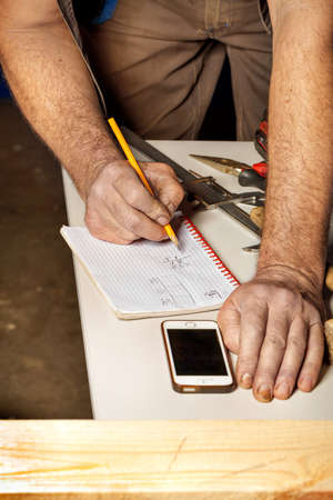 The men's hard-working hands of the carpenter write down the necessary calculations in a notebook with a pencil for subsequent work. Blurred tool background. Foreground in blur for copy space. Vertical image.の写真素材