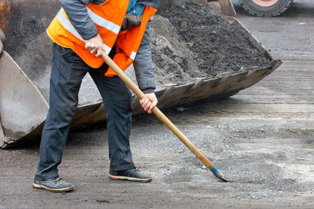 A road worker in a bright orange vest prepares an old surface for repairs and removes road debris in...