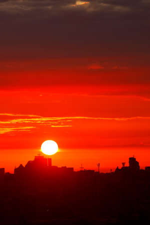 The bright sun in the early morning floods the sky with red light, rising above the silhouettes of the houses of the sleeping city. Vertical image, copy space.の写真素材