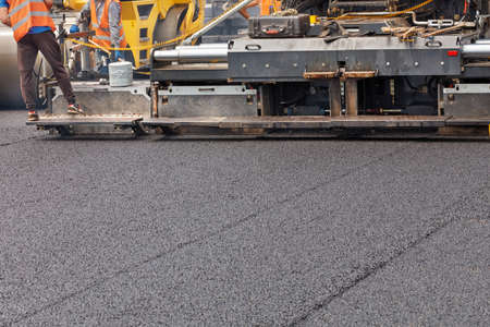 An industrial paver is laying a layer of new hot asphalt on the roadway. Workers control the...