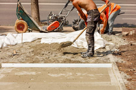 A worker levels the foundation with sand and a shovel under a wooden level for laying paving slabs...