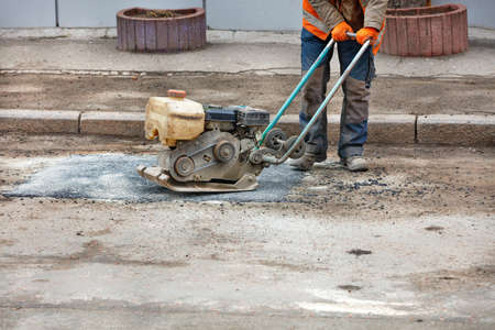A worker with an old vibrating plate for road construction repairs potholes on an old road.
