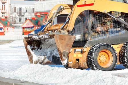 The metal bucket of a compact grader clears snow from a town square on a sunny winter day.の写真素材