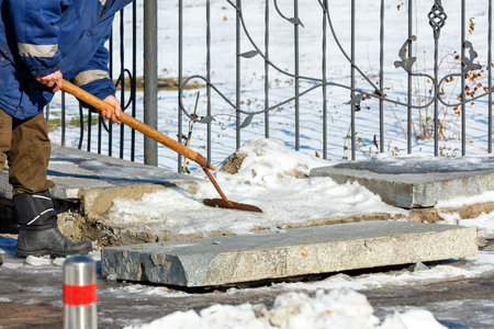 On a clear winter day, a utility worker clears snow and ice from a granite curb on a city street.の写真素材