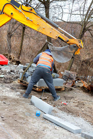A construction worker using a road excavator bucket unloads concrete curbs to build a walkway....