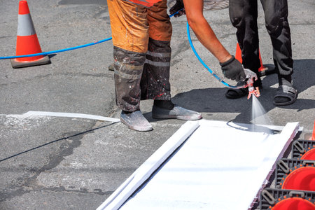 A road worker in dirty overalls applies white road markings to the asphalt pavement of the road with a spray gun on a summer day. Copy space.の写真素材