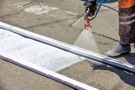 A road worker in dirty overalls applies white road markings with a spray gun on a summer day. Copy...