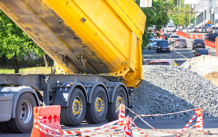 Crushed stone is unloaded from the body of a large dump truck against the backdrop of a city street on a sunny summer day. Copy space.の写真素材