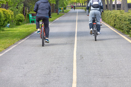 Two young men ride bicycles on an asphalt path among the green trees of a city park on a sunny day....