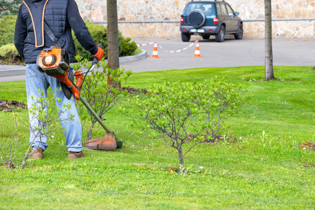 A gardener with a trimmer carefully takes care of the green lawn of a city park while walking...