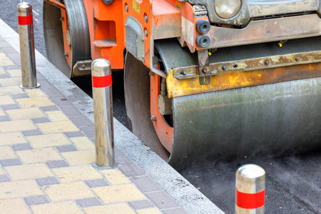 The metal cylinder of a vibrating roller compacts fresh asphalt on a sunny day on the roadway....