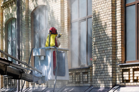 A worker in a protective suit, standing in a construction telescopic cradle, cleans the dirty coating from the brick wall of the facade of the house with a sandblaster.の写真素材