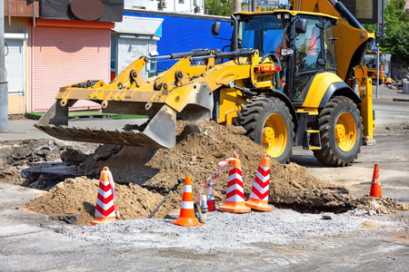 A large yellow backhoe excavator digs into the ground, surrounded by construction cones and debris, as workers focus on repairing the street in a bustling urban area.の写真素材
