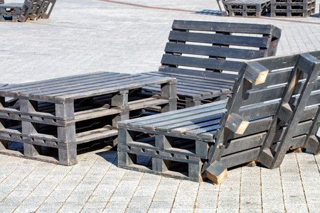 In a lively urban plaza, a unique seating area features creatively arranged wooden pallets turned into chairs and tables. The bright sunlight casts playful shadows on the paved ground.の写真素材
